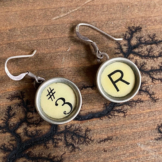 Pair of earrings with 'R' and '#3' on round yellow stones, on a wooden surface.
