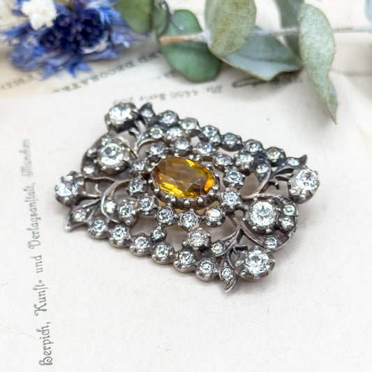 Decorative brooch with central yellow gemstone and surrounding crystals on a textured surface with floral elements.