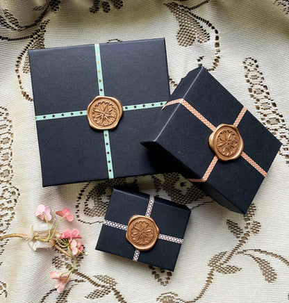 Three navy gift boxes tied with patterned ribbon and stamped starburst wax seals, staged on lace fabric with dried pink blossoms, showing the packaging rather than the earrings.