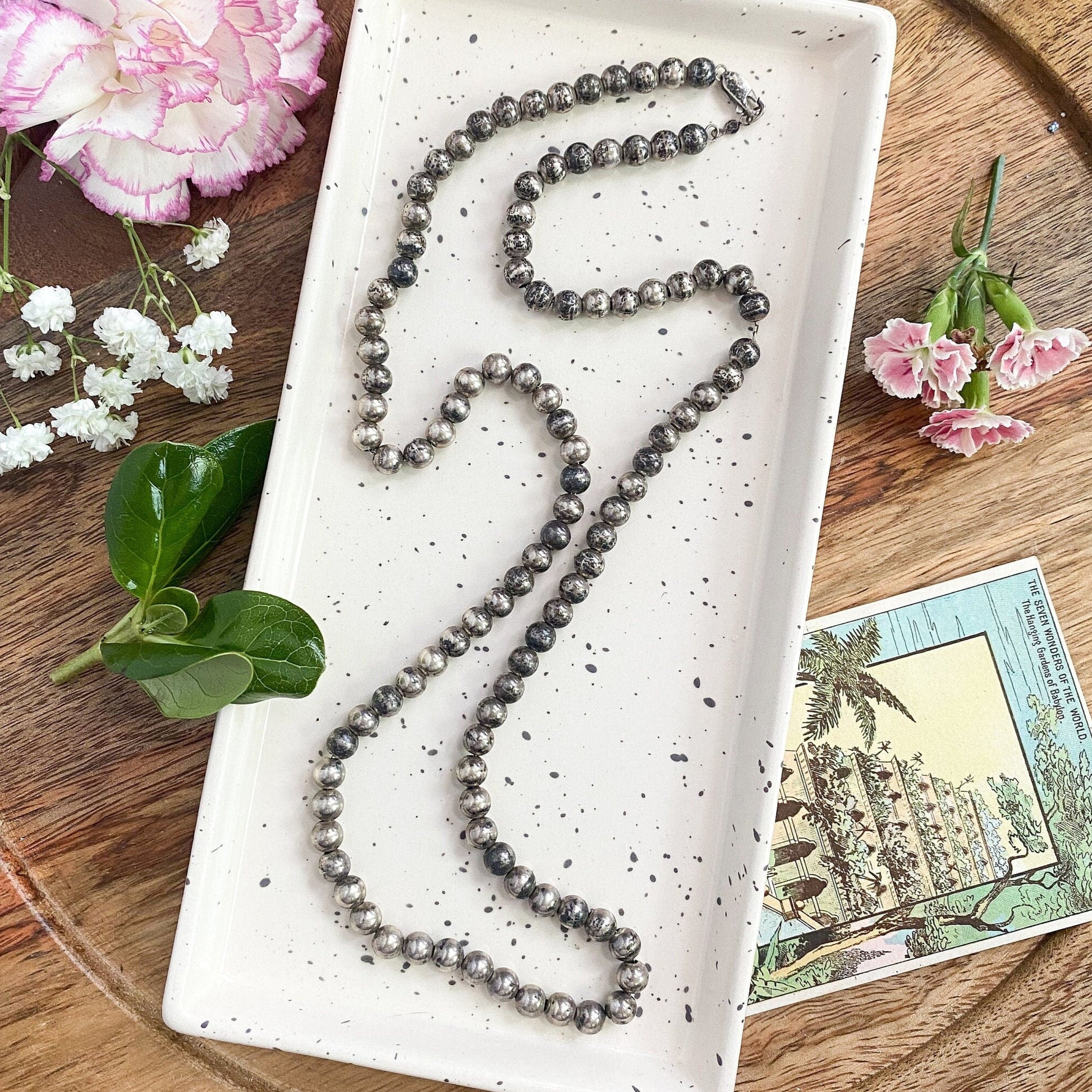 Necklace on a white dish with flowers and a postcard on a wooden surface