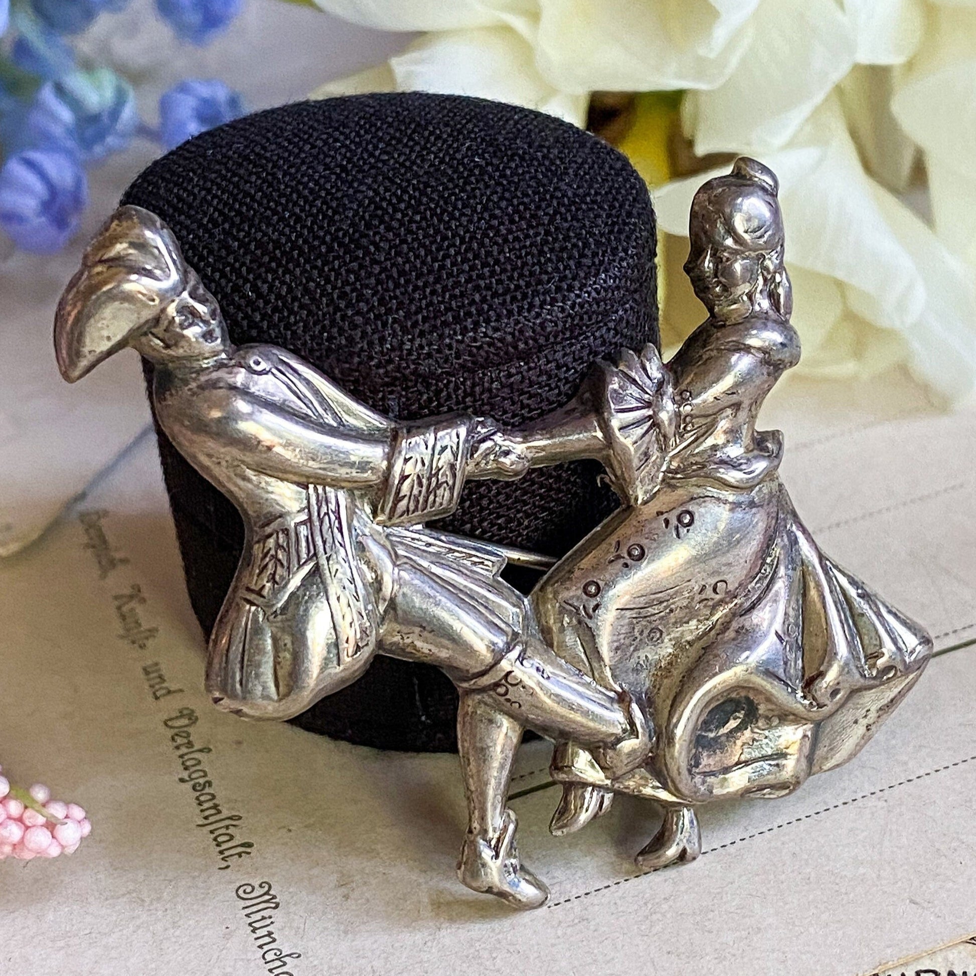 Silver figurine of two people embracing on a black cushion with flowers in the background