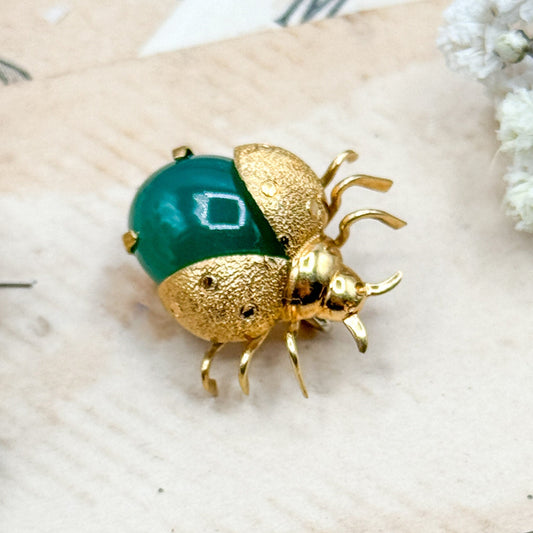 Gold ladybug brooch with green gem on a textured surface with flowers
