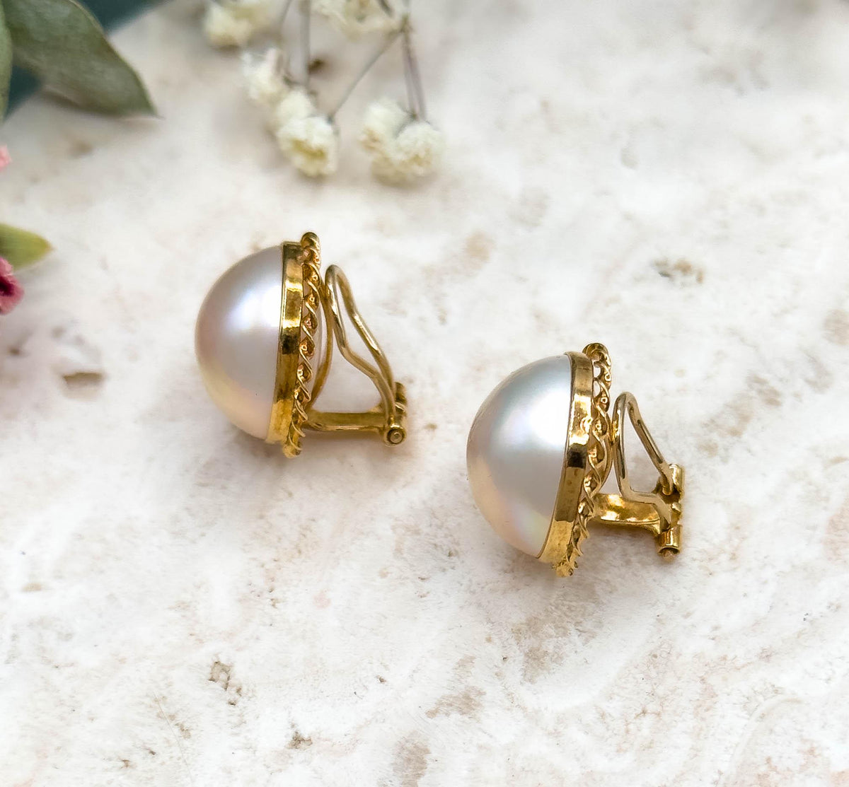 Three-quarter view from behind showing the hinged yellow gold clip fittings standing out from each domed pearl, with the rope twist bezel edge curving away on the stone slab.
