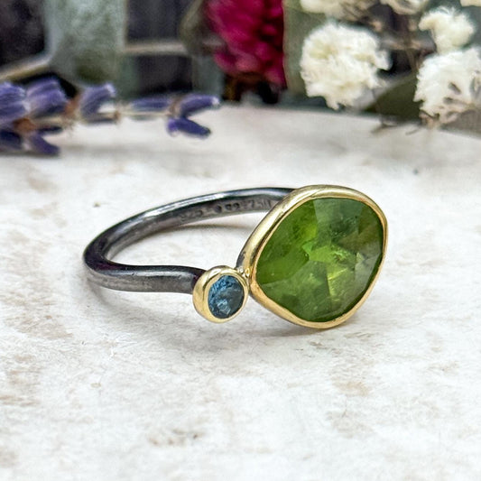 Ring with green gemstone and blue stone on a textured surface with flowers in the background