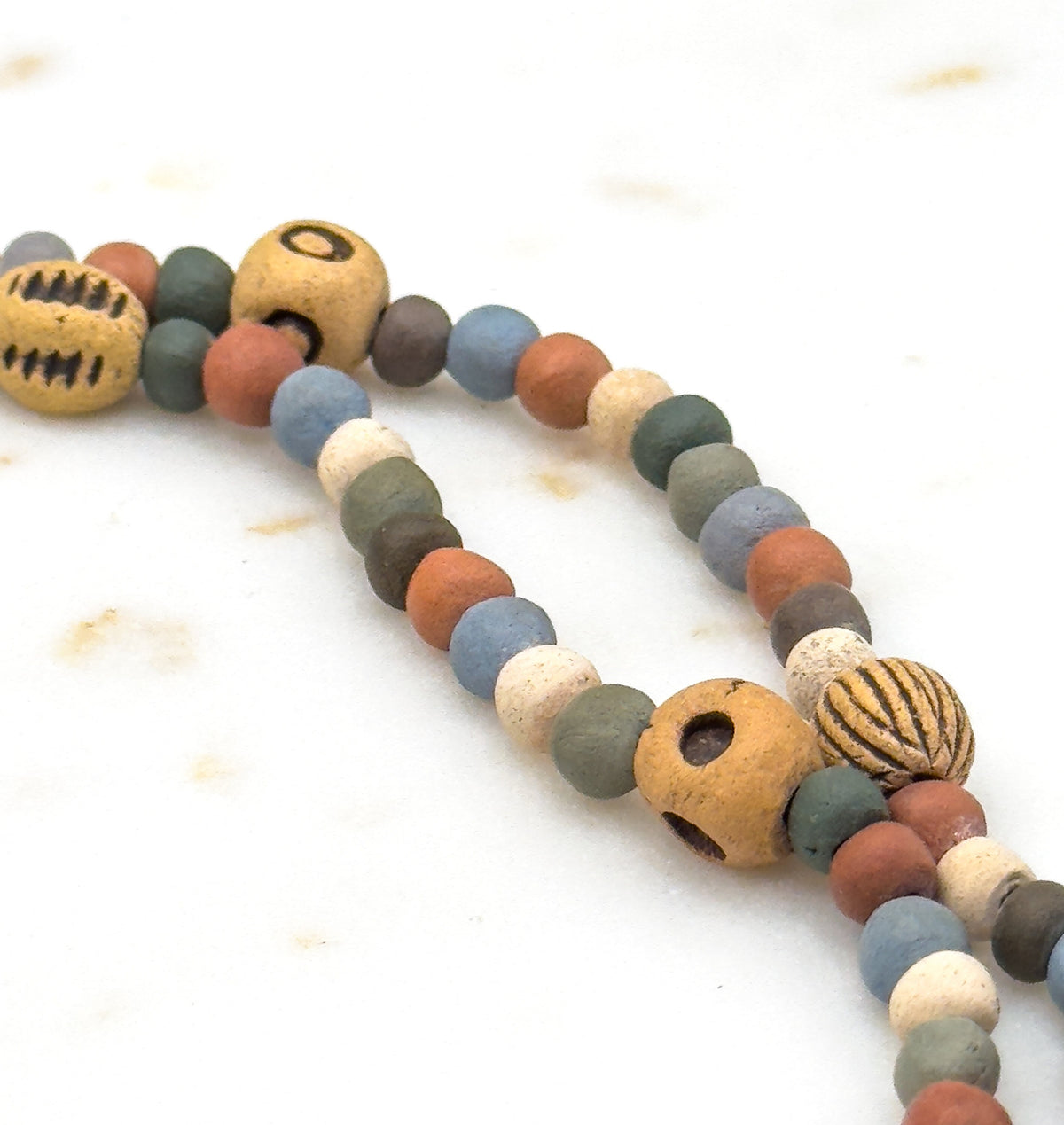 Close crop on a looping curve of the strand against pale marble, where a big ochre bead shows two wide drilled holes beside a ribbed pumpkin-shaped bead and rows of small muted rounds.