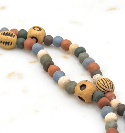 Close crop on a looping curve of the strand against pale marble, where a big ochre bead shows two wide drilled holes beside a ribbed pumpkin-shaped bead and rows of small muted rounds.