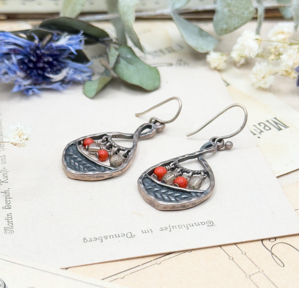 Three-quarter side view on the printed cards, showing the thickness of the hammered teardrop edges, the small ball ends of the ear wires and the beads swinging inside each frame.