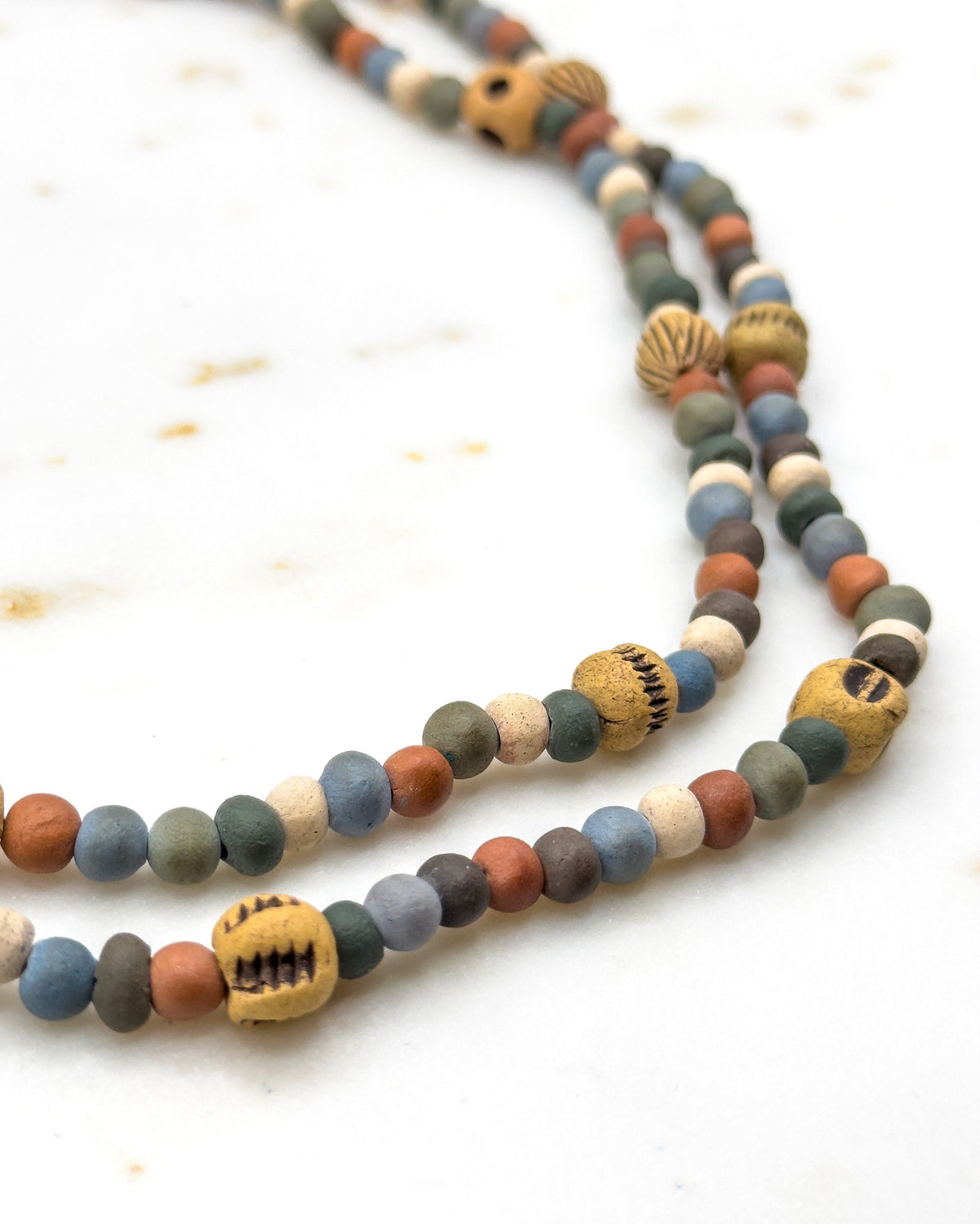 Two doubled strands curving side by side across pale marble, the near beads sharp and the far ones softening into blur, with notched ochre beads sitting at intervals along each row.