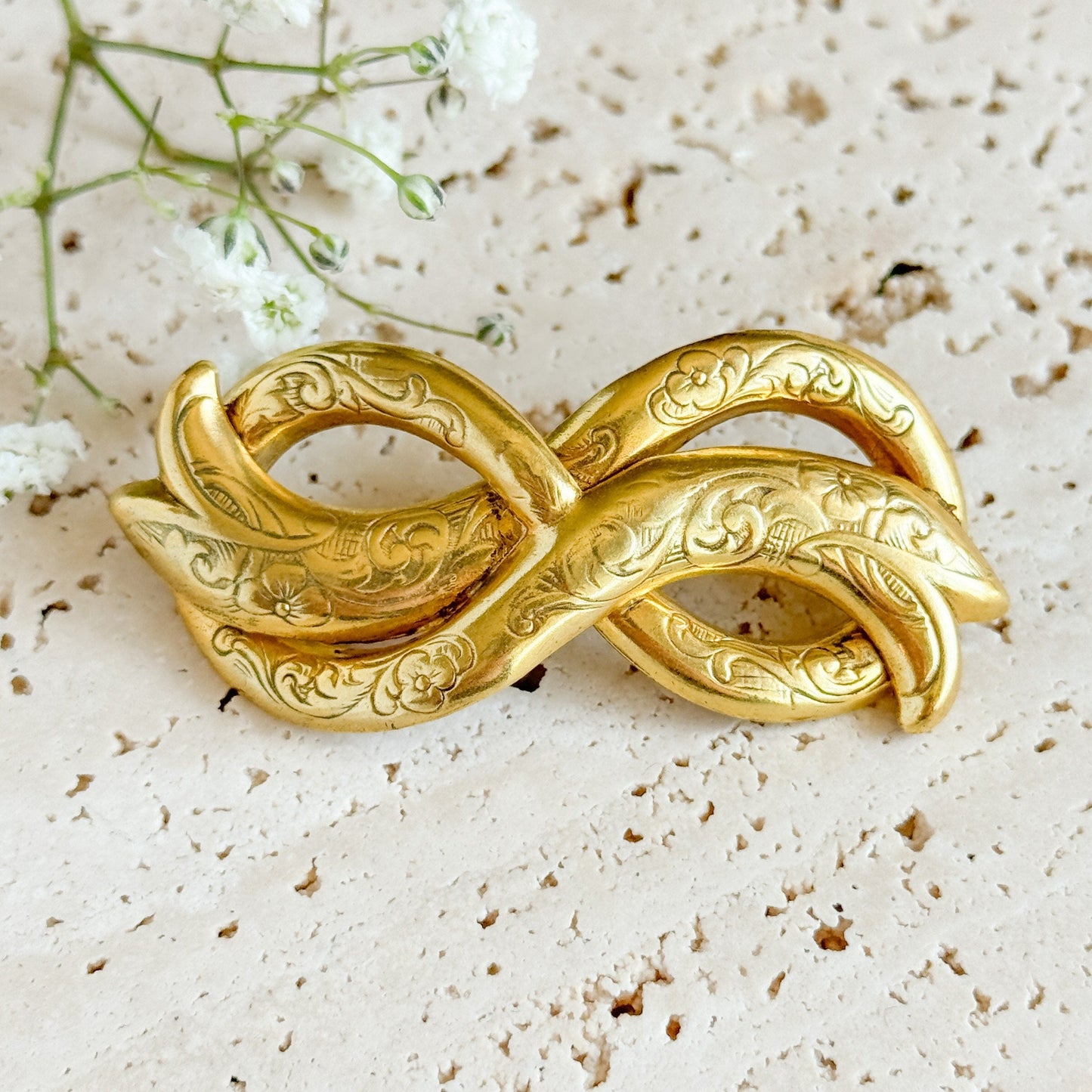 Gold infinity symbol brooch on a textured surface with small flowers.