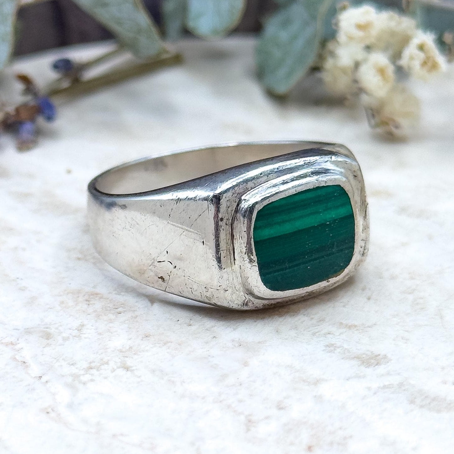 Silver ring with a green stone on a textured surface with plants in the background