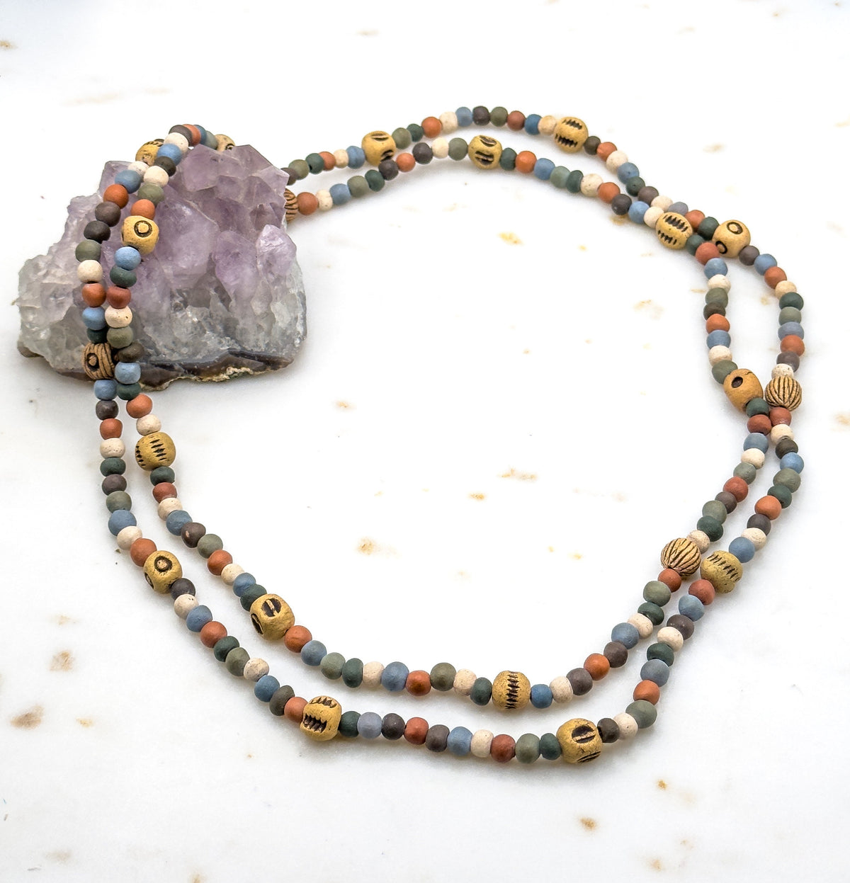 The full necklace coiled in a double loop on a white speckled surface, one end draped over a pale purple rock, showing the small round clay beads and scattered larger patterned ones.