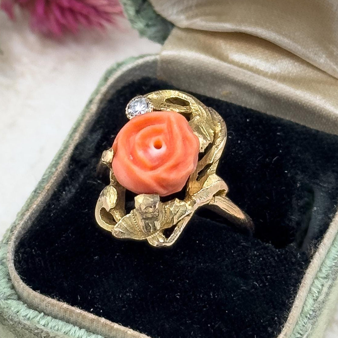Gold ring with coral-colored stone in a green velvet box on a textured surface.