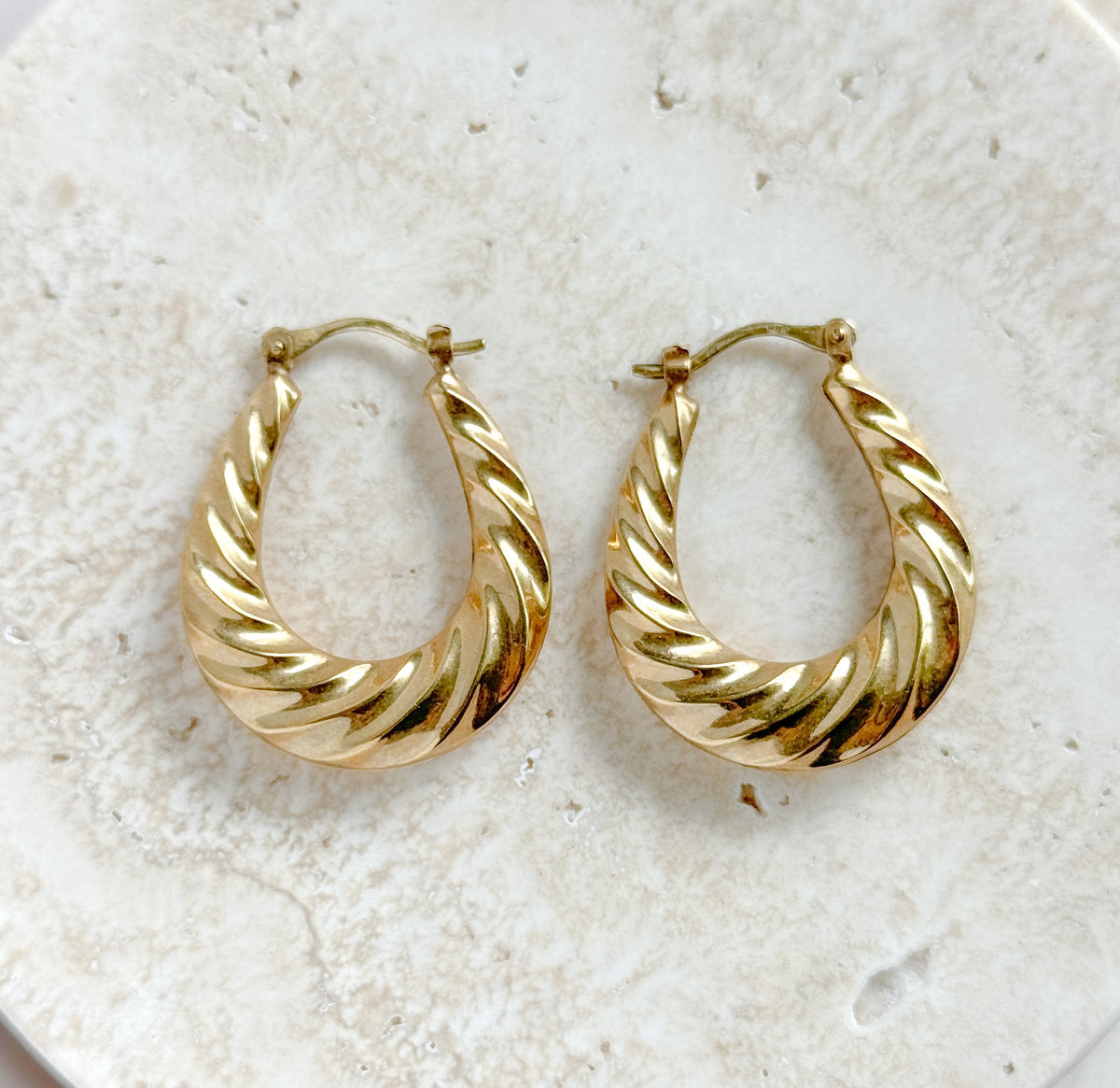 Both hoops laid side by side on a pale speckled stone surface, front faces up, showing the twisted ribbed gold and the hinged wire catches at the top.