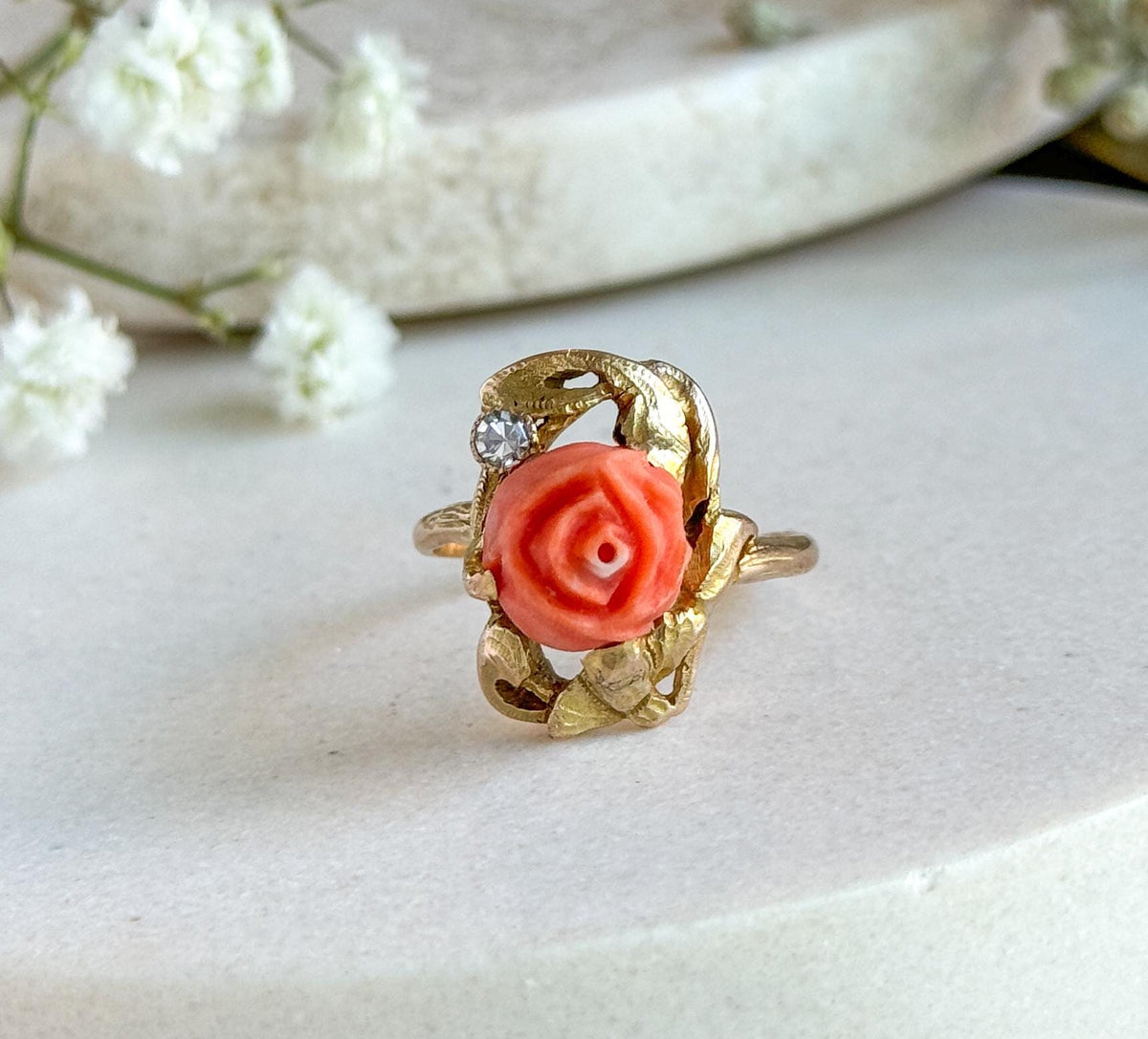 Head-on close crop of the face, filling the frame, showing the spiralled petals of the carved coral rose, the small bezel-set diamond above it and the gold leaves curling around, on pale stone.