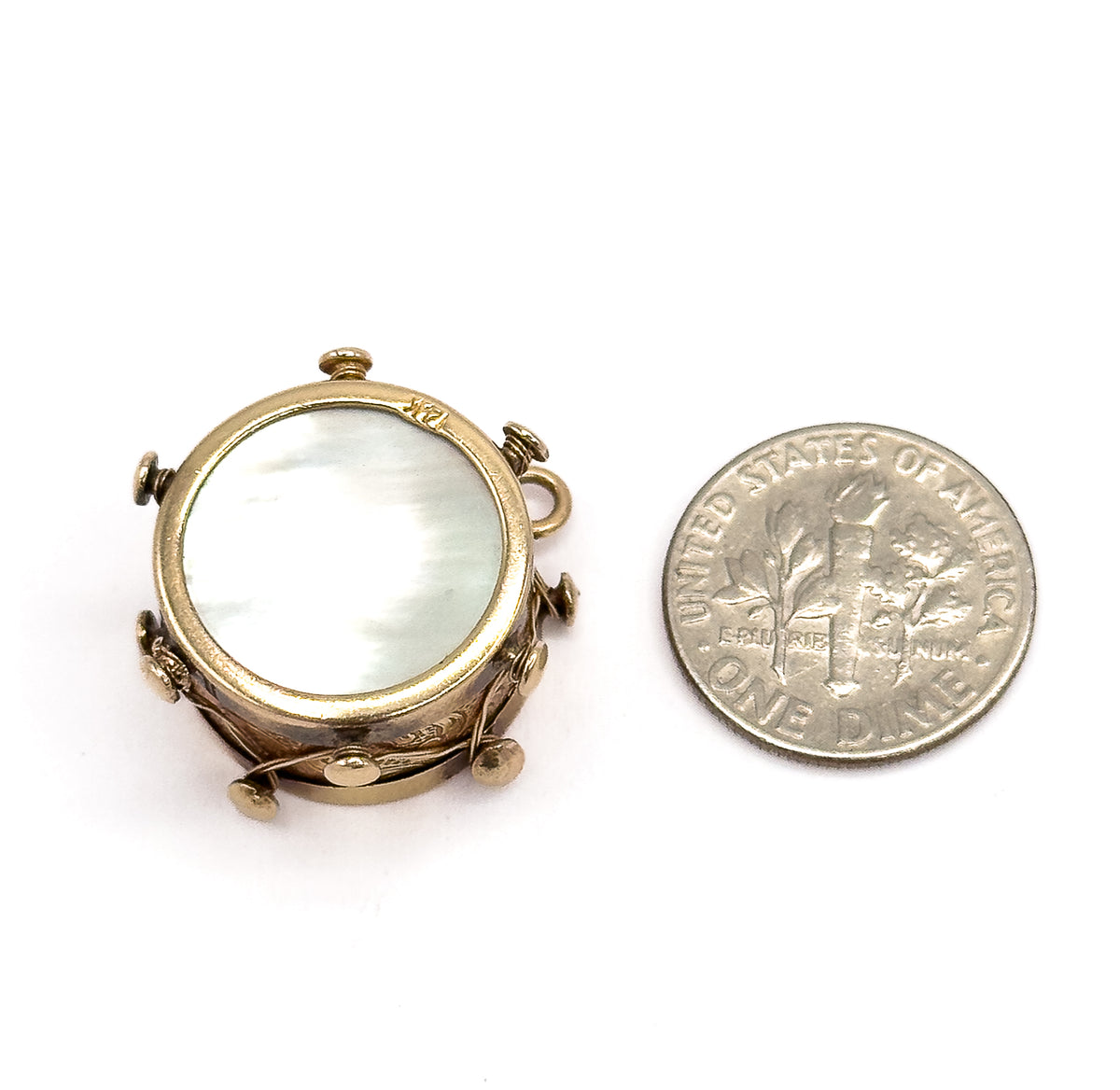 Placed beside a dime for scale, the drum charm shows its mother of pearl face and knobbed rim tilted toward the viewer, the small bail projecting at the right.