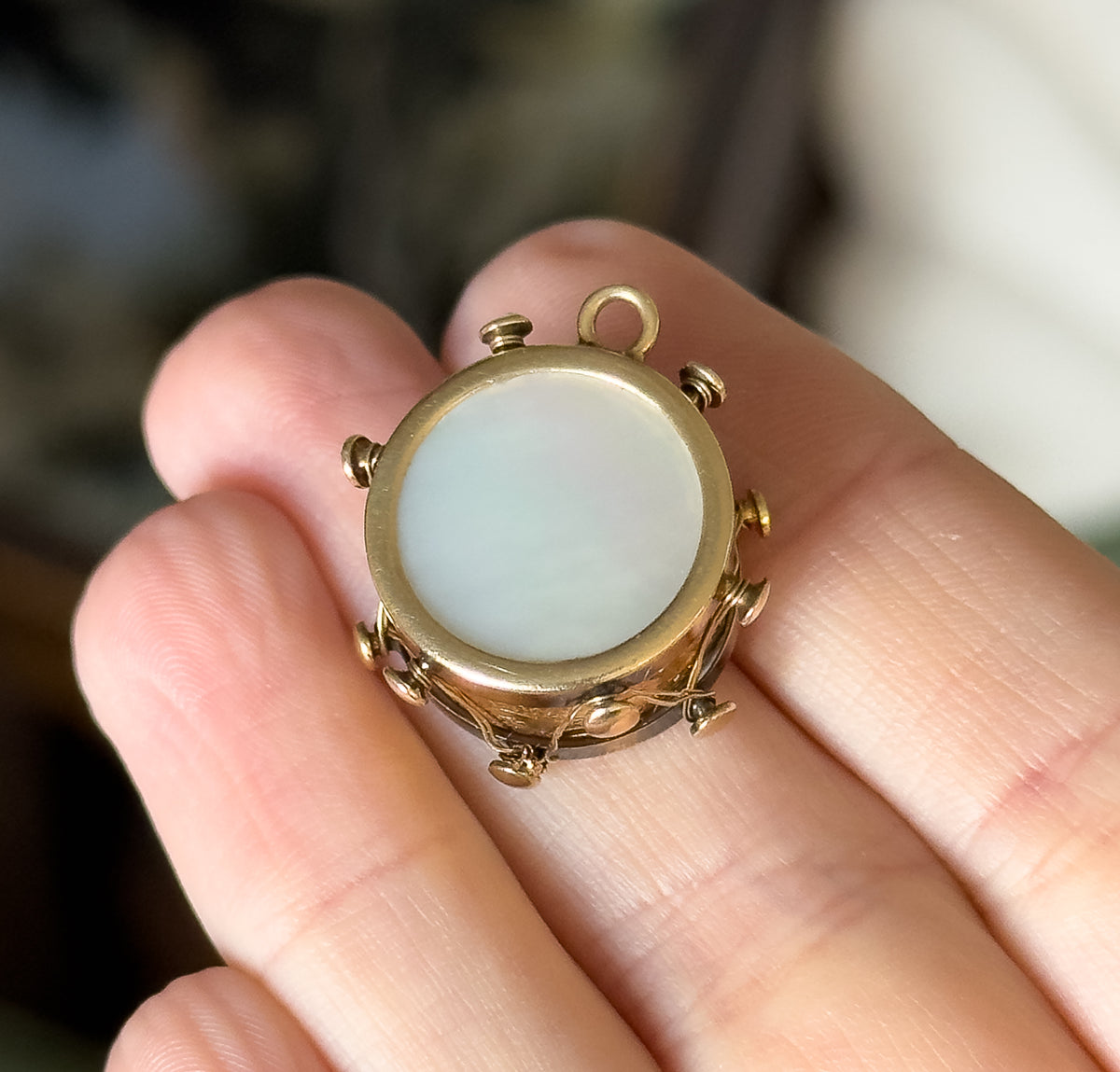A vintage 14 karat gold drum charm with a pale iridescent mother of pearl face and small bail, shown resting on fingertips that give a sense of its small size.