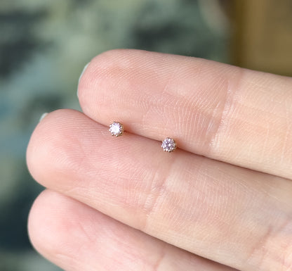 The two round pink glass studs rest on a fingertip, their prong settings and 14K yellow gold bezels showing how tiny each stone is against the skin.