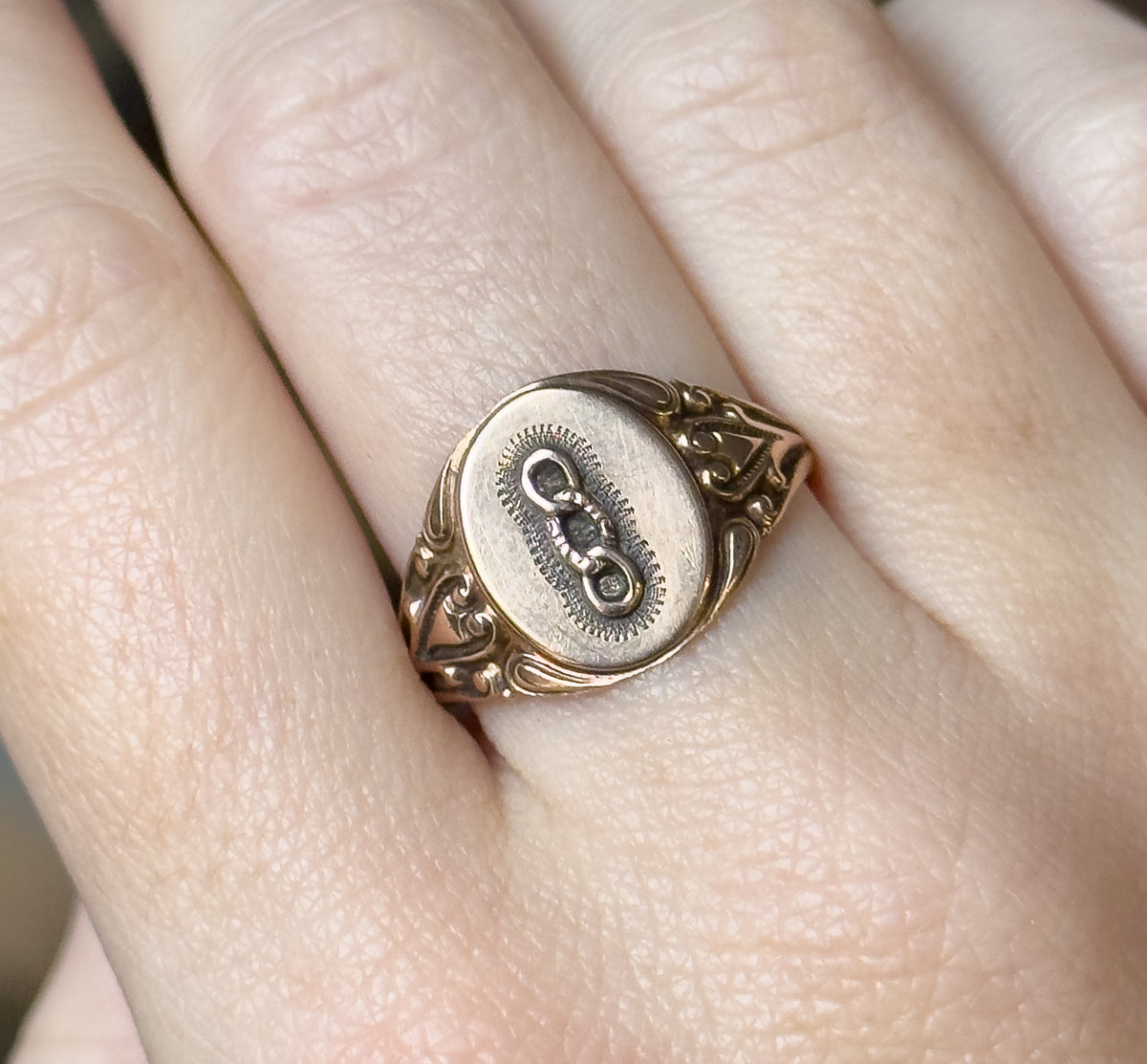Close view on the finger showing the three-link emblem raised within a finely textured oval border, with the scrolled shoulders curving down either side of the face.