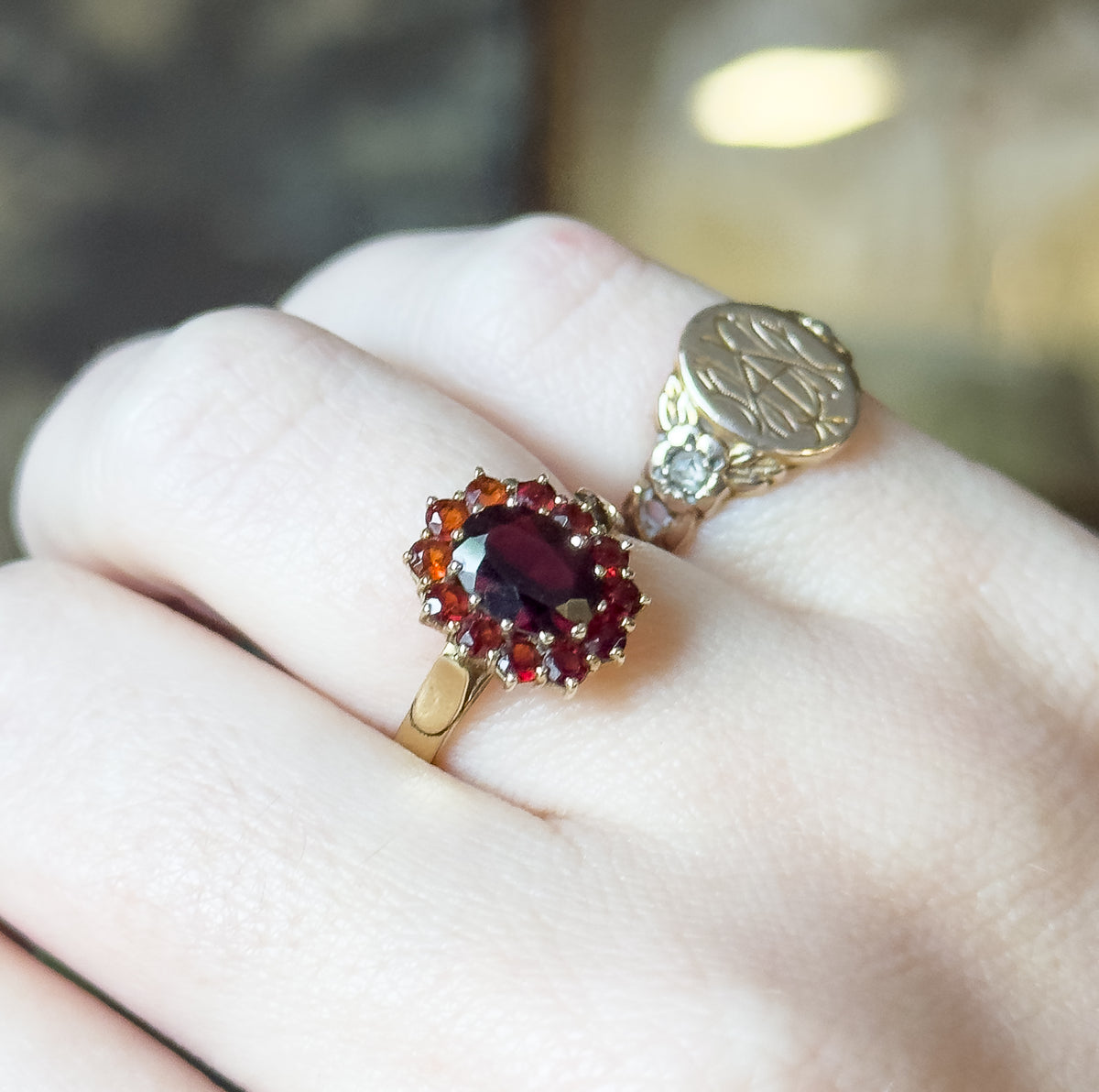 Angled close-up on the hand, the garnet cluster sitting high on one finger beside another ring with an engraved oval face, the fingers giving a clear sense of its scale.