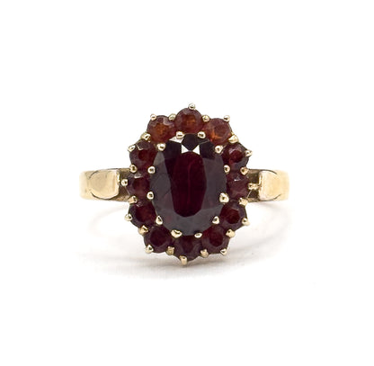 Face-on view against white with the ring upright, the oval garnet centred in its prong-set bezel inside a full ring of twelve small round garnets and squared yellow gold shoulders either side.