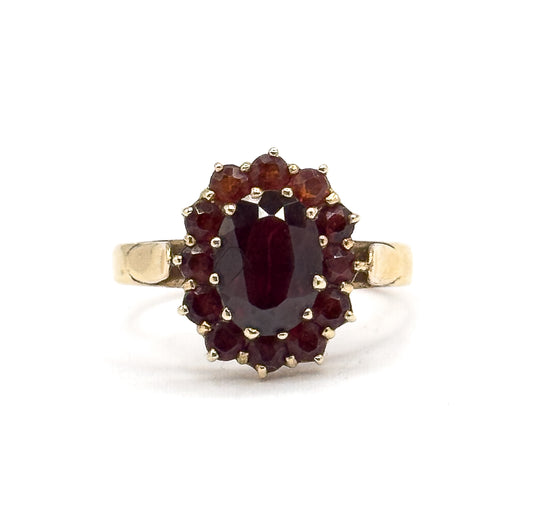 Face-on view against white with the ring upright, the oval garnet centred in its prong-set bezel inside a full ring of twelve small round garnets and squared yellow gold shoulders either side.