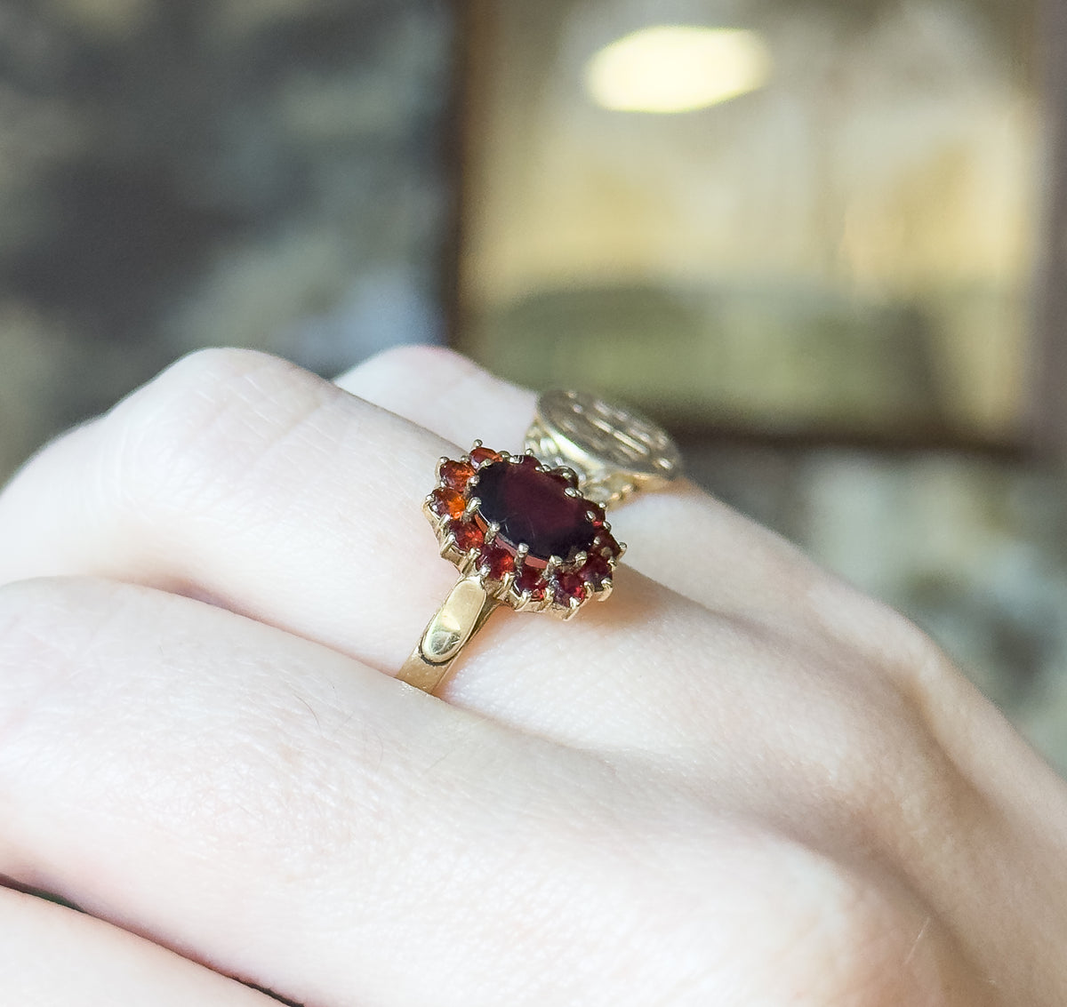 A garnet cluster ring in 14K yellow gold, shown on a finger, its oval garnet ringed by twelve small prong-set garnets above a tapering shank.