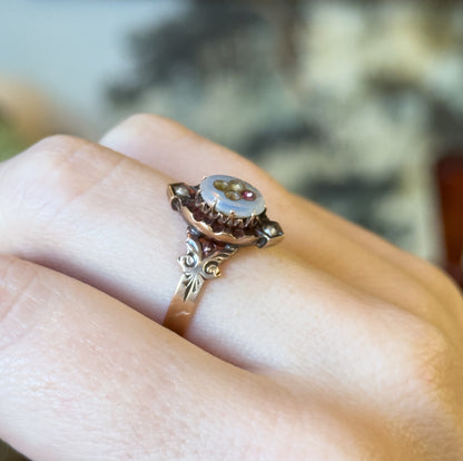Victorian 14K yellow gold ring with a milky glass paste oval face, pink and colourless glass inlay and seed pearl accents, shown on a finger with engraved shoulders in profile.