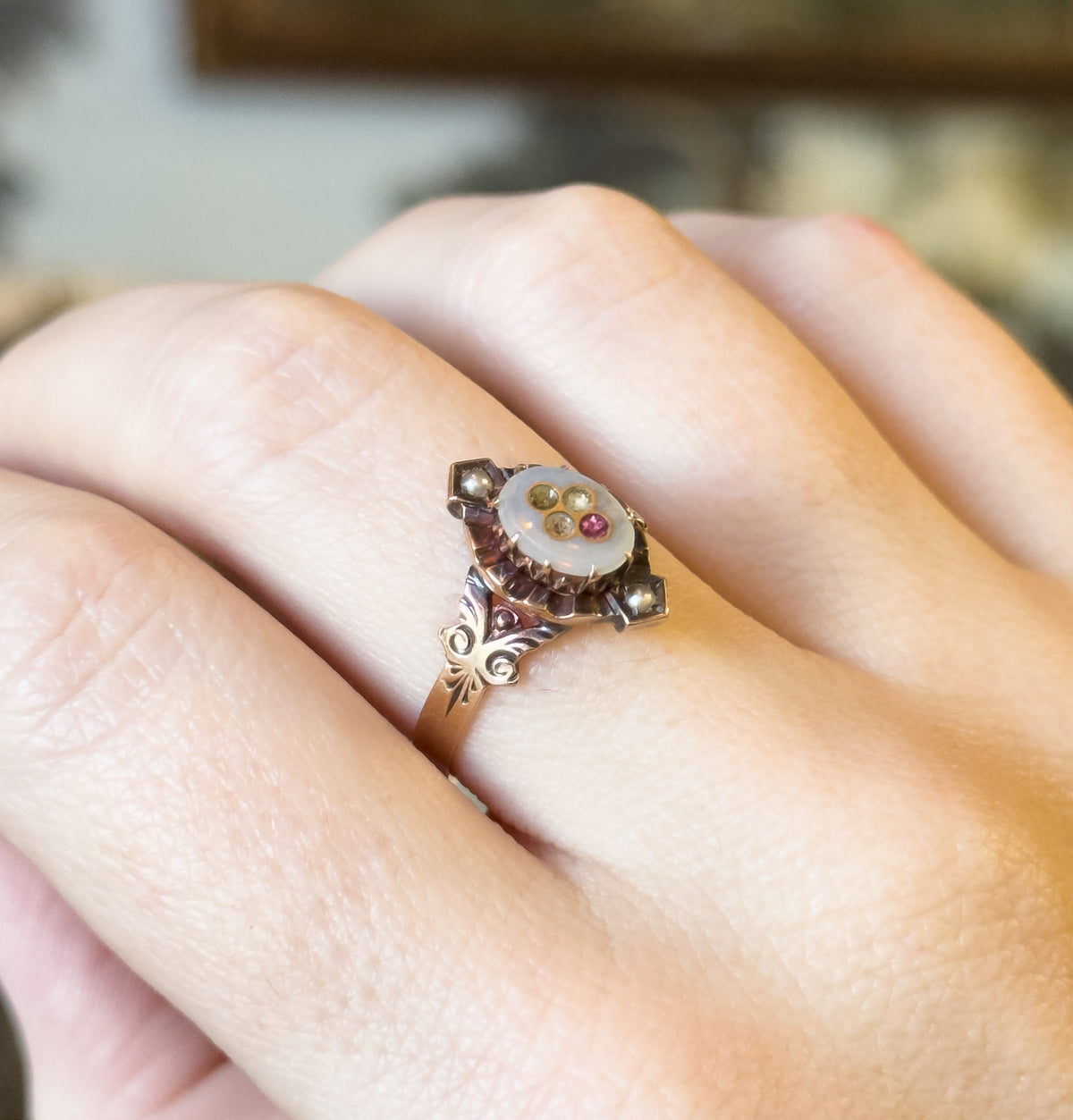Shown on a finger from above, the oval face tilts toward the camera so the pink glass inlay, seed pearls and scrolled engraved shoulders meet the narrow shank.