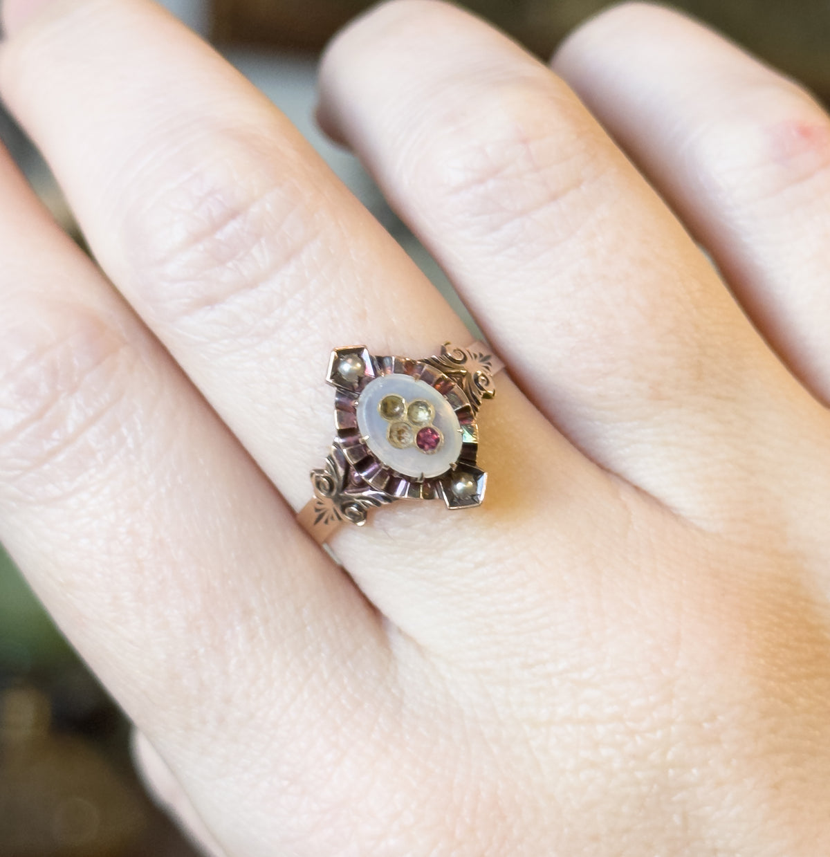 Close view across the hand, the ring's oval face filling the space between two fingers so its four-point gallery, corner seed pearls and tapering shank are seen against skin for scale.