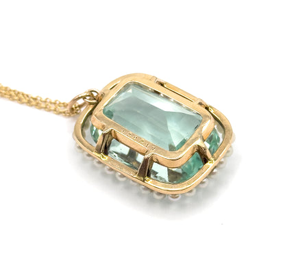 Close view of the underside, the aquamarine's pavilion glowing green through the open gold framework while the seed pearls peek out along the rim and the chain trails off to the left.
