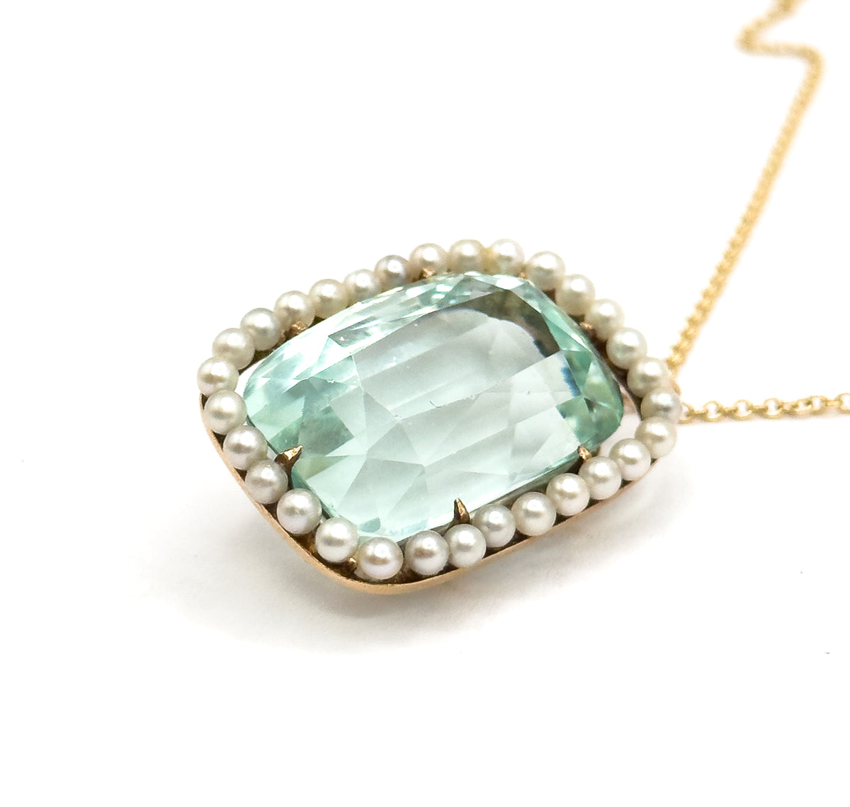 Tilted face-up view on white, the seafoam aquamarine's broad facets held by small gold prongs inside the full ring of seed pearls, with the yellow gold chain sweeping off to the upper right.