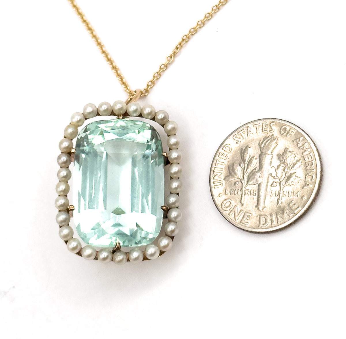 Face-up on white beside a US dime for scale, the pendant only slightly taller than the coin, its seafoam aquamarine and seed pearl halo above the yellow gold chain rising to the top edge.