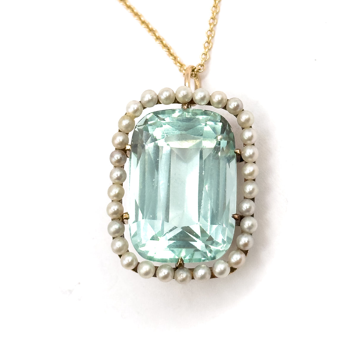 Straight-on and close, the cushion aquamarine's stepped facets fill the frame within the even ring of seed pearls, gold prongs gripping each corner and the chain rising centrally from the bail.
