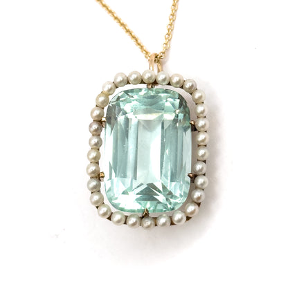 Straight-on and close, the cushion aquamarine's stepped facets fill the frame within the even ring of seed pearls, gold prongs gripping each corner and the chain rising centrally from the bail.