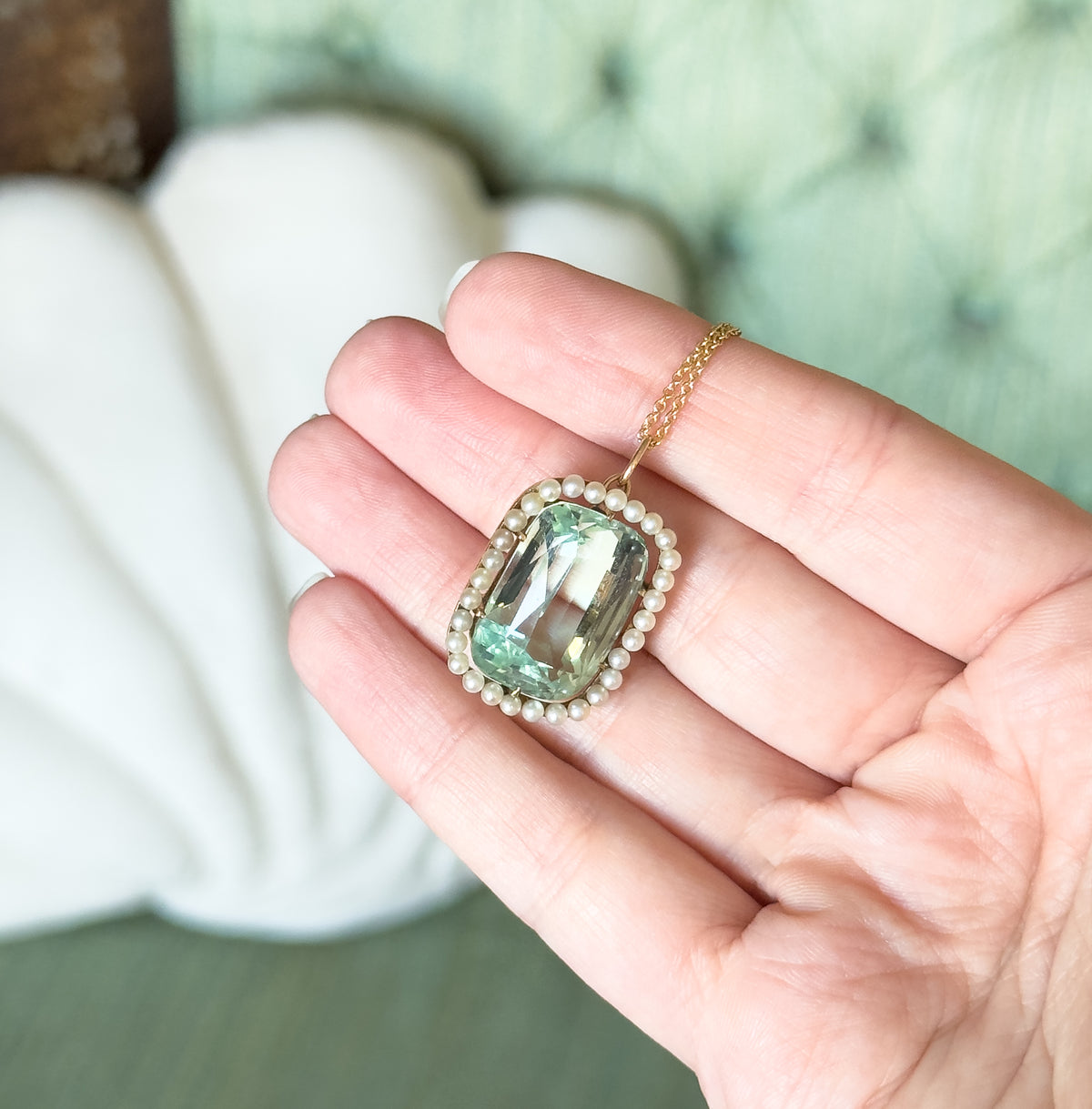 The pendant rests on open fingers, giving a sense of scale: the seafoam aquamarine and its seed pearl halo span roughly a fingertip's width, with the gold chain draped over the hand.