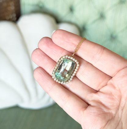 The pendant rests on open fingers, giving a sense of scale: the seafoam aquamarine and its seed pearl halo span roughly a fingertip's width, with the gold chain draped over the hand.
