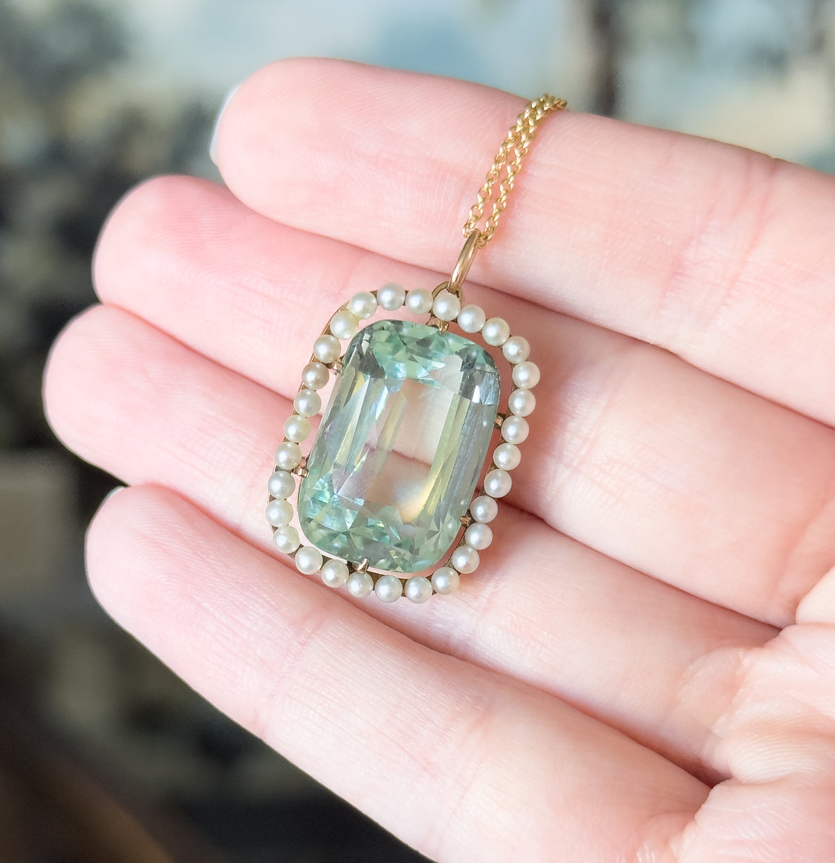 Closer still on the same hand, the seafoam aquamarine glowing through its basket setting with gold prongs at the corners and the bail carrying the chain up between two fingers.