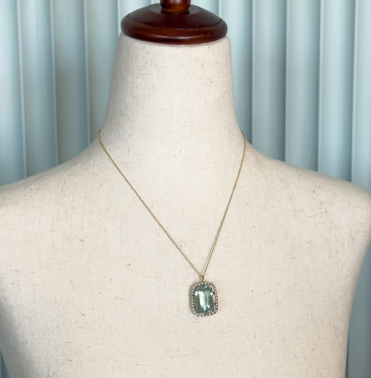 Shown at the neck on a linen dress form, the fine gold chain tracing the collarbone with the seafoam aquamarine pendant and its pearl halo hanging flat below.