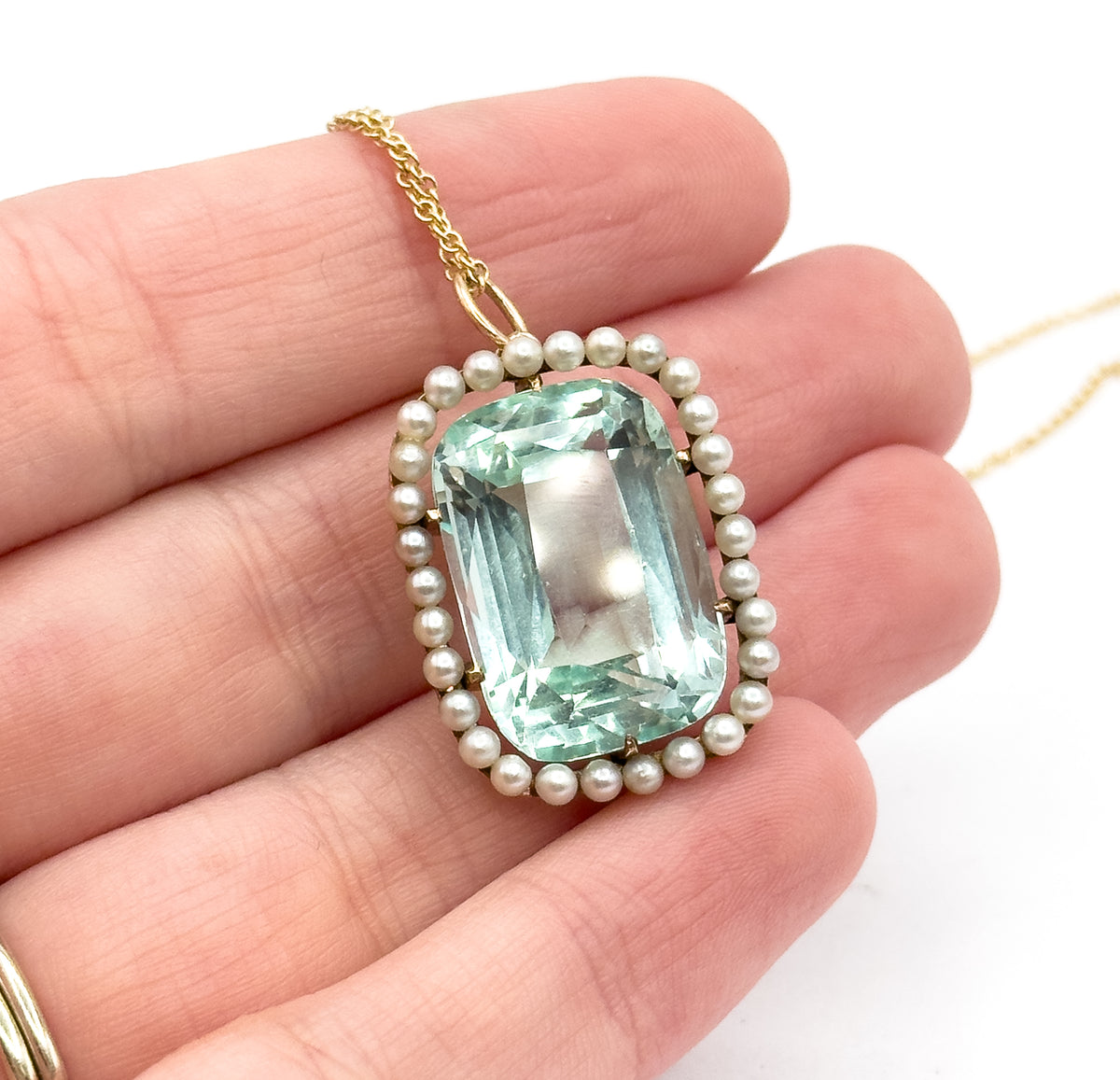 The pendant lies across the middle fingers at an angle, the pearl halo tilted so the round beads catch the light one by one around the aquamarine's edge.