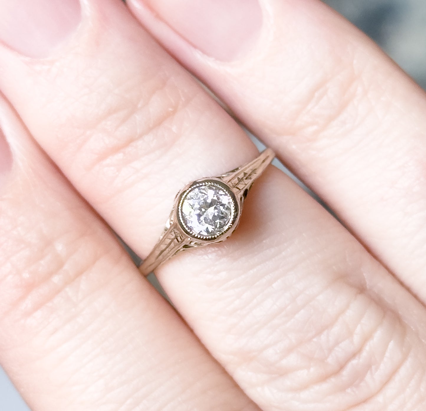 Shown on a finger in close crop, giving scale — the bezel-set diamond covers little more than a third of the finger's width, with the engraved flower shoulders and split shank visible from above.