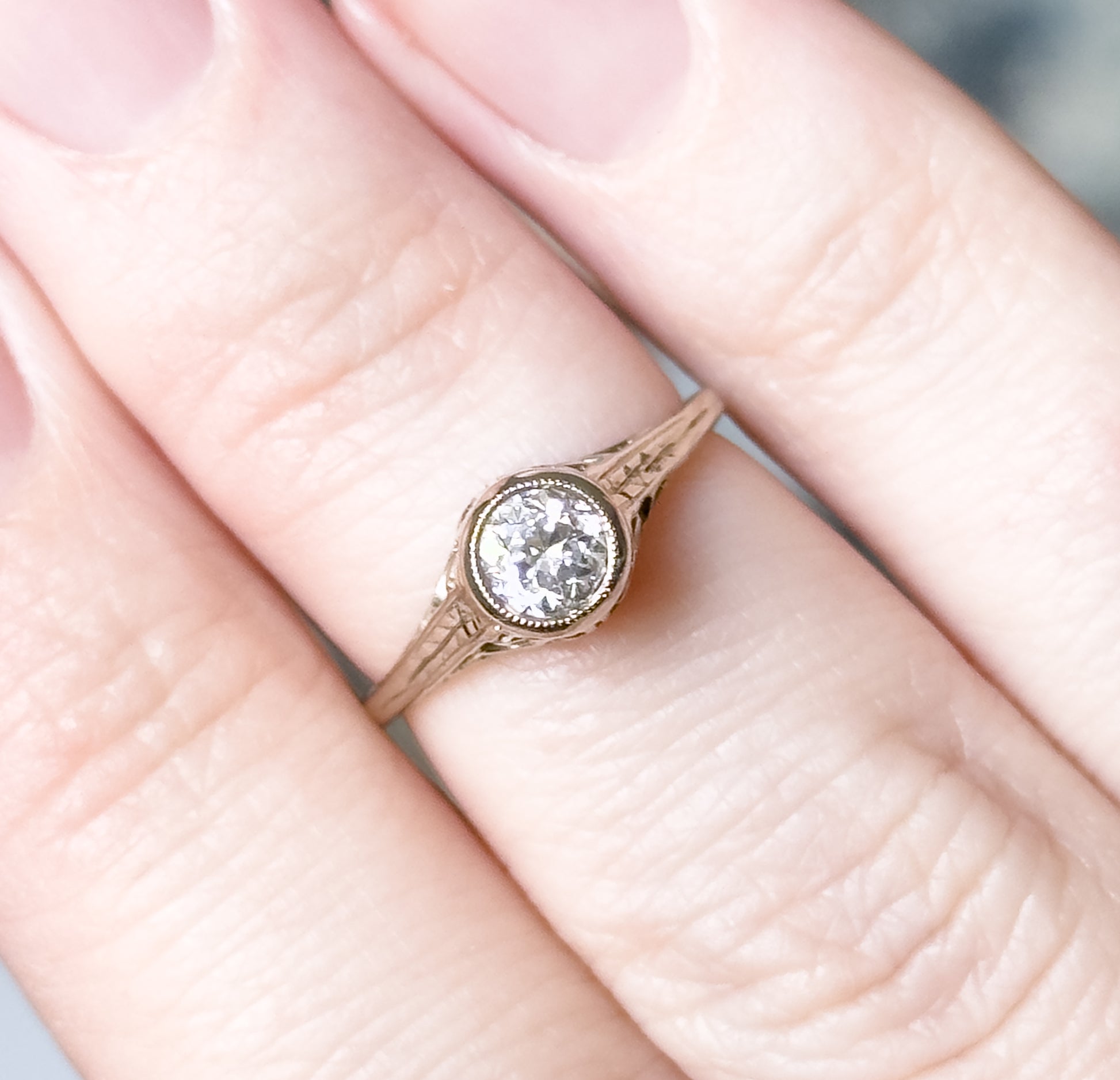 Shown on a finger in close crop, giving scale — the bezel-set diamond covers little more than a third of the finger's width, with the engraved flower shoulders and split shank visible from above.