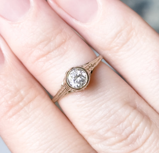 Shown on a finger in close crop, giving scale — the bezel-set diamond covers little more than a third of the finger's width, with the engraved flower shoulders and split shank visible from above.