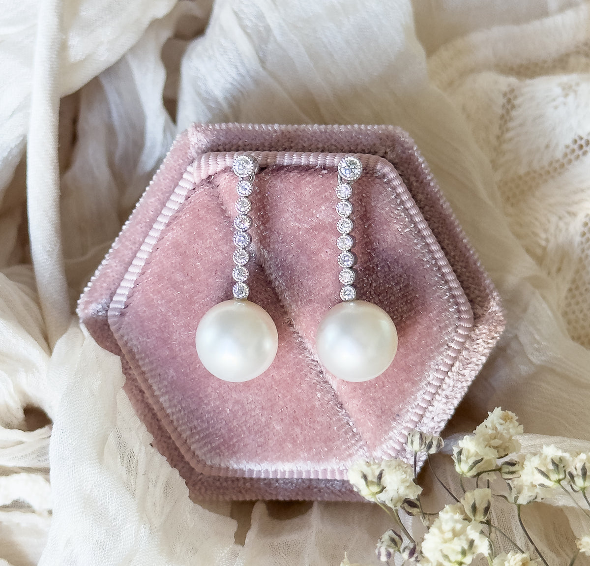 Cultured pearl and diamond drop earrings in platinum, shown upright on a pink velvet hexagonal box, each round pearl hanging below a vertical line of bezel-set round diamonds.