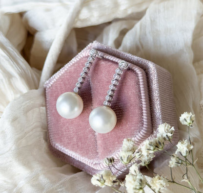 Angled overhead view on the pink velvet box amid cream fabric and dried white blossoms, showing the graduated diamond line tapering down to each creamy pearl drop.