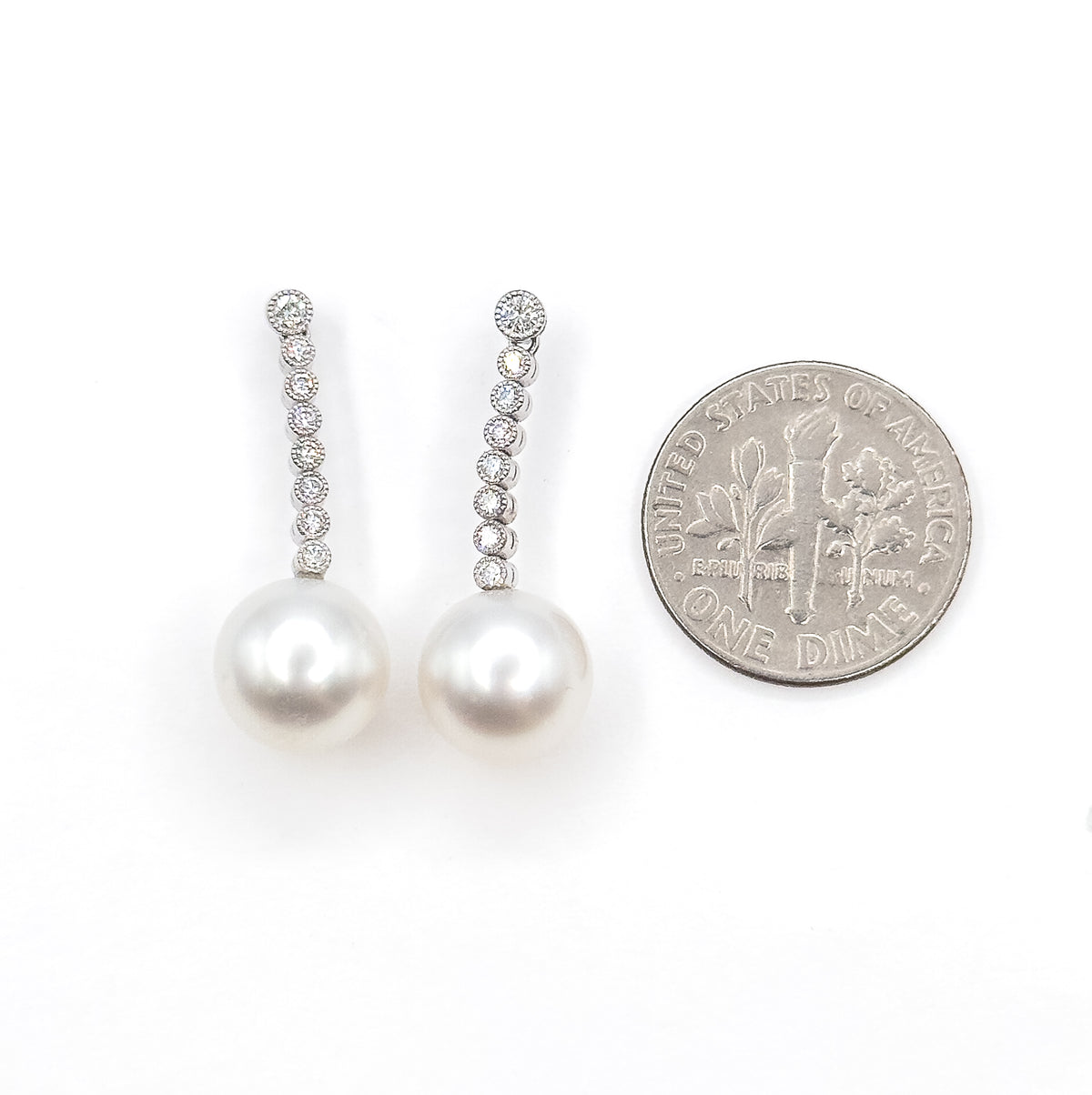 Both earrings laid flat on white next to a US dime, the coin standing about as tall as one graduated diamond line, with each creamy pearl drop below roughly two-thirds the coin's width.