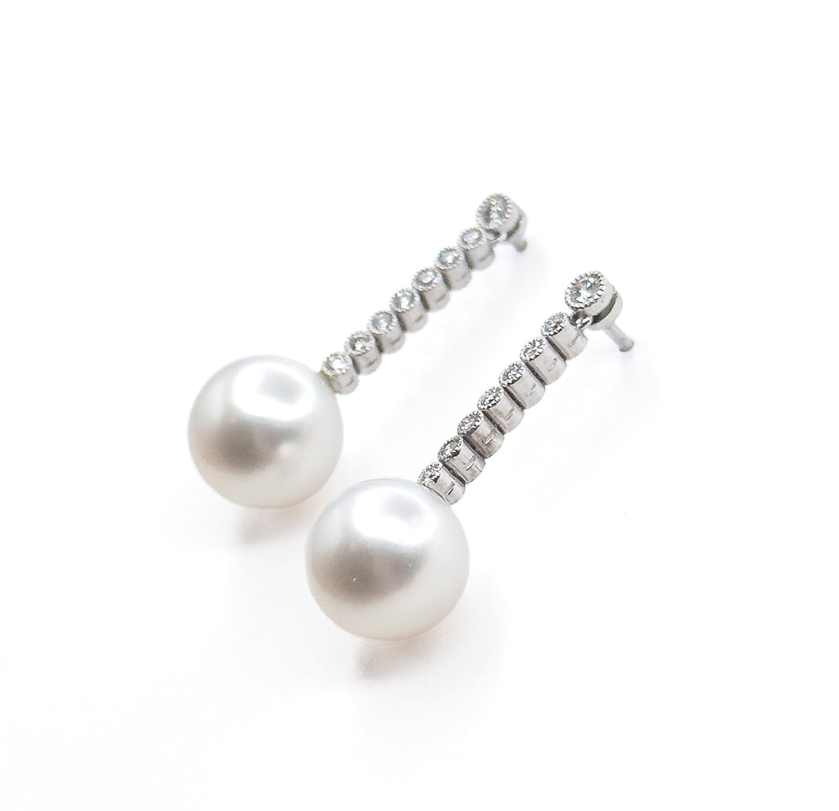 Both earrings on plain white, tipped diagonally so the light catches each bezel-set diamond along the graduated lines and the soft sheen curving across the two creamy pearl drops.