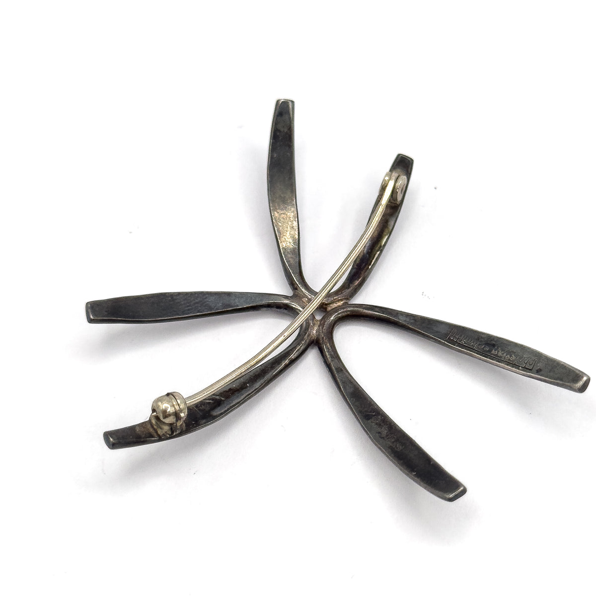 Full back view on white, the pin stem stretched diagonally across the radiating rays from its coiled hinge at one tip to the catch at another, with stamps faintly visible.