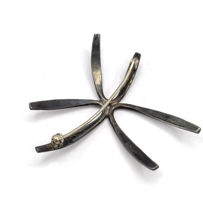 Full back view on white, the pin stem stretched diagonally across the radiating rays from its coiled hinge at one tip to the catch at another, with stamps faintly visible.