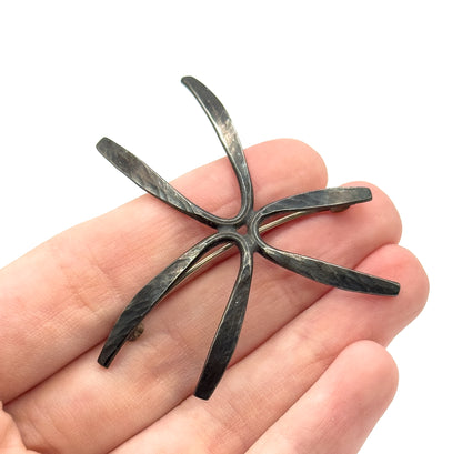 Mid-century hammered starburst brooch in sterling silver, shown resting on an open palm with its six tapering rays spread wide, dark patina settled into the hammered texture, pin stem visible beneath.