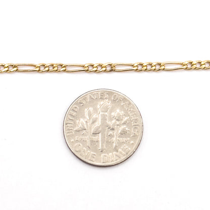 A straight length of the Figaro chain laid above a dime, the coin showing how the alternating round and long links measure only a fraction of its width.